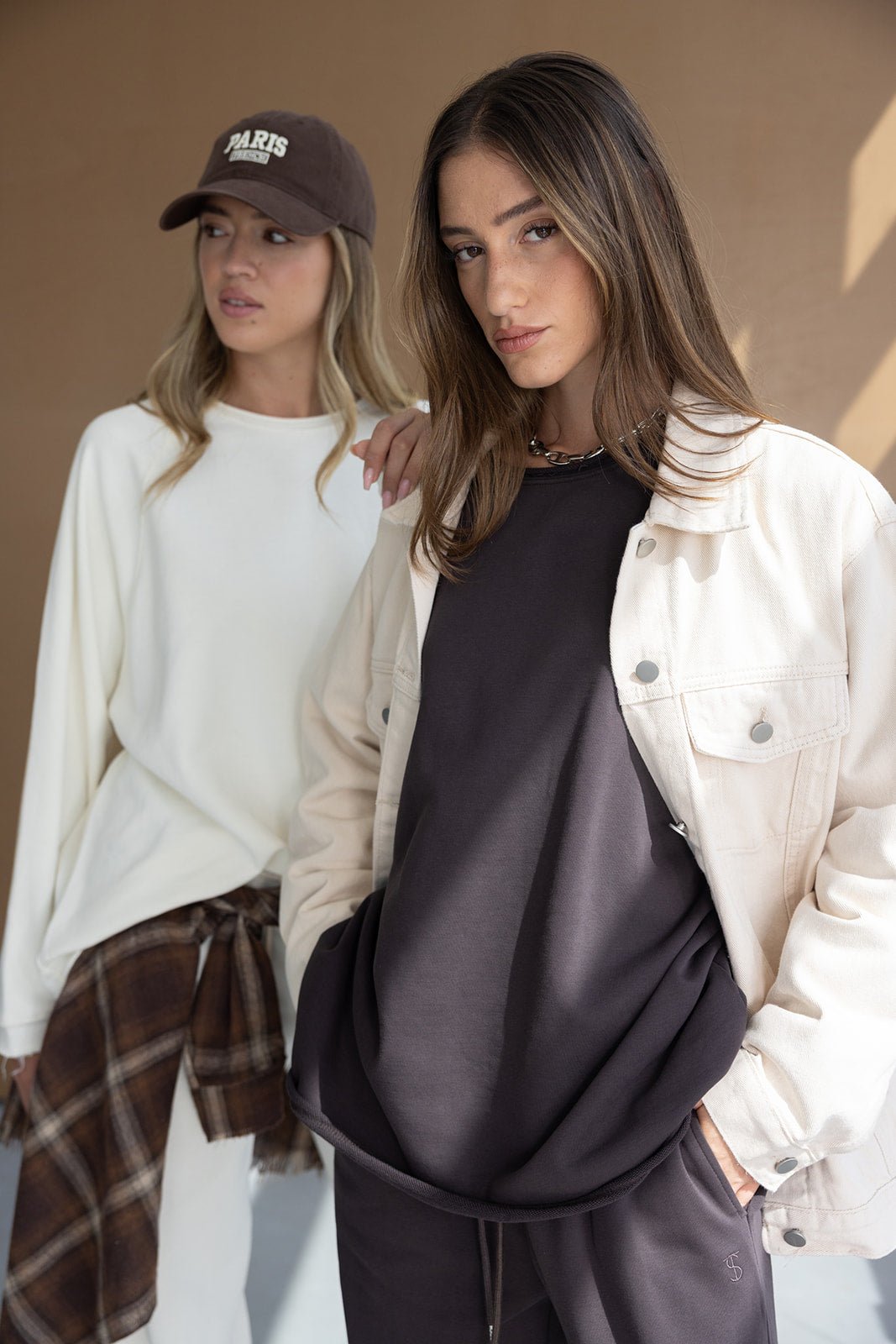 שתי נשים לובשות בגדים אופנתיים בסגנון קז'ואל לנוער, אחת עם ג'קט בז' ומכנסיים כהים, השנייה בלבן וכובע מצחיה