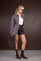 אישה לבושה בחולצת פלאנל משובצת, גופייה לבנה, מכנסי עור קצרים, תיק טרנדי ונעלי עקב חומות, אופנה עדכנית לנשים