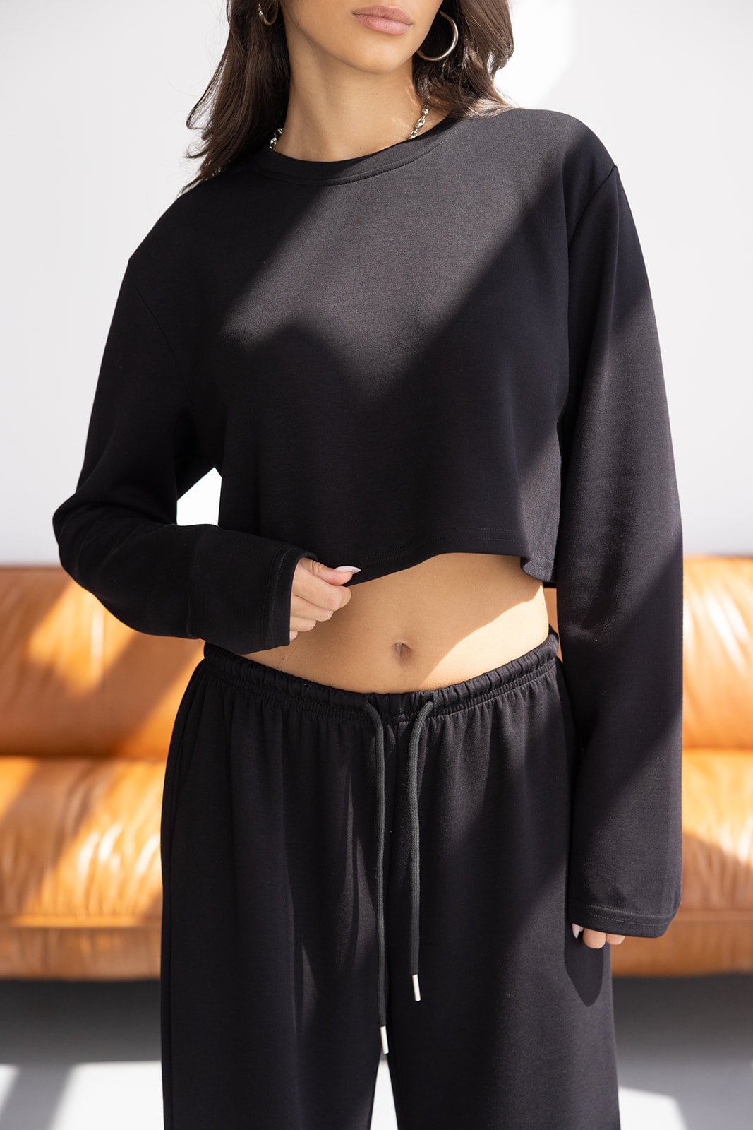 אישה לובשת סט טרנינג שחור אופנתי עם חולצה קצרה ומכנסיים, רקע ספה חומה