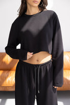 אישה לובשת סט טרנינג שחור אופנתי עם חולצה קצרה ומכנסיים, רקע ספה חומה