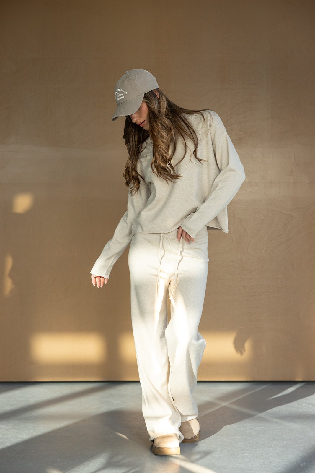 דוגמנית בלבוש קז'ואל אופנתי לנשים, סווטשירט ומכנסיים בצבע קרם, כובע מצחייה ונעלי בית