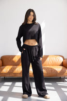 דוגמנית לובשת סט טרנינג שחור לנשים בגזרת קרופ ומכנסיים רחבים, עומדת ליד ספה חומה בסלון מואר