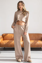 צעירה לובשת סט טרנינג נשים אופנתי בצבע בז', חולצה קרופ ארוכה ומכנסיים רחבים, על רקע ספה חומה