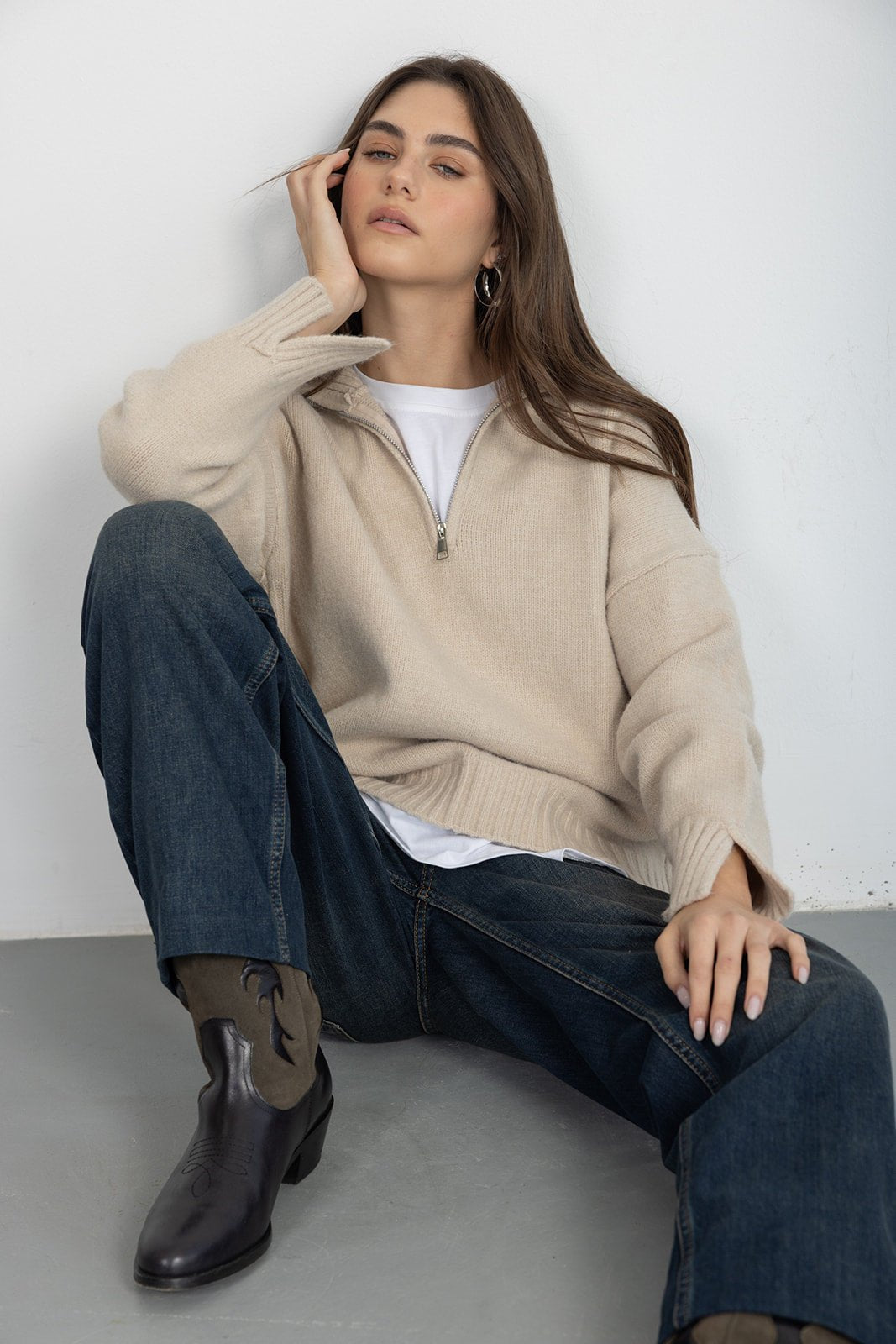 דוגמנית עם סוודר בז' עם רוכסן, ג'ינס כחול ומגפיים אופנתיים יושבת על רצפה בסגנון קז'ואל