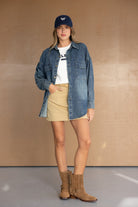 אישה עם חולצת טי לבנה, חצאית מיני בז', חולצת ג'ינס, מגפוני זמש חומים וכובע כחול, לוק קז'ואל אופנתי לנשים