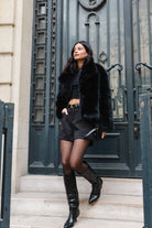 אישה לובשת ג'קט פרווה שחור, טופ קצר, מכנסיים קצרים, גרבונים ומגפיים אופנתיים, עומדת בכניסה עירונית אופנתית