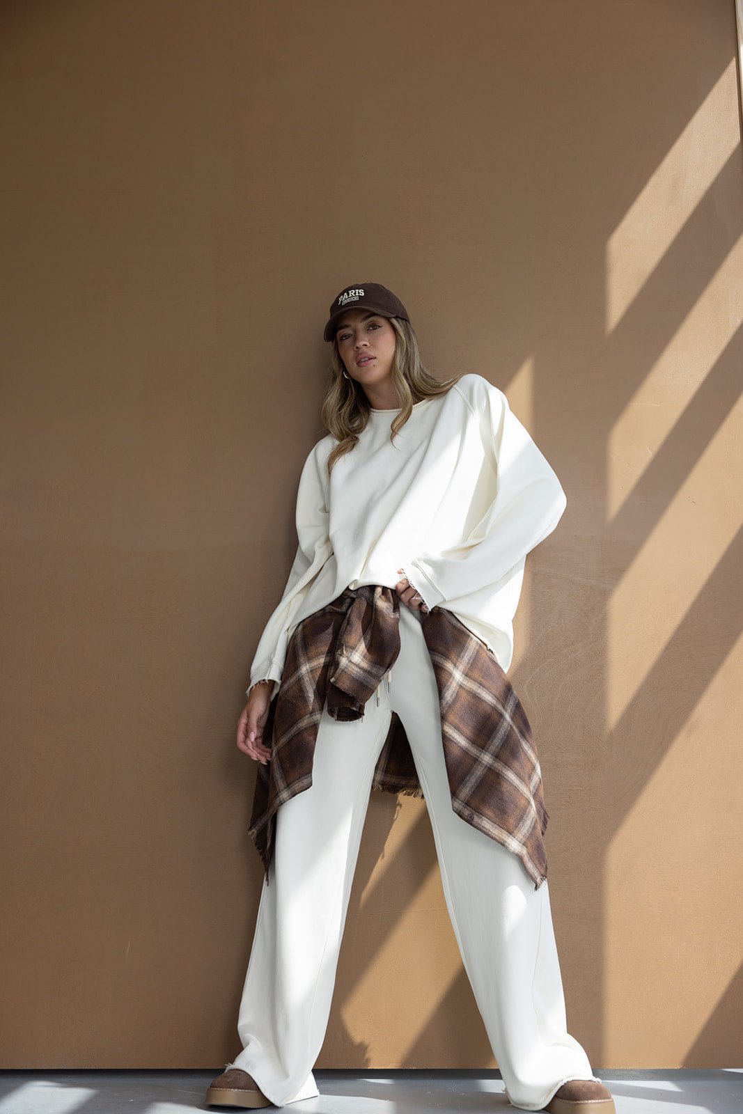 דוגמנית לובשת סט בגדי נשים לבן עם חולצה ומכנסיים רחבים, חולצה משובצת קשורה למותן וכובע קז'ואל חום