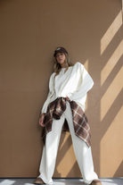 דוגמנית לובשת סט בגדי נשים לבן עם חולצה ומכנסיים רחבים, חולצה משובצת קשורה למותן וכובע קז'ואל חום