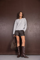 דוגמנית לובשת סוודר אפור, מכנסי עור קצרים, מגפי בוקרים חומים ותיק מעוצב. אופנת נשים טרנדית.