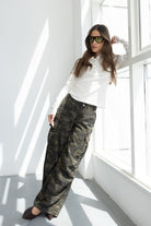 דוגמנית לובשת חולצה לבנה עם שרוול ארוך, מכנסיים רחבים עם הדפס קמופלאז' ומשקפי שמש ירוקים, בסטודיו מואר