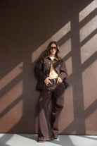 אישה לובשת חליפת ג'ינס חומה, ג'קט ומכנסיים רחבים, משקפי שמש, מצטלמת מול קיר חום באור טבעי