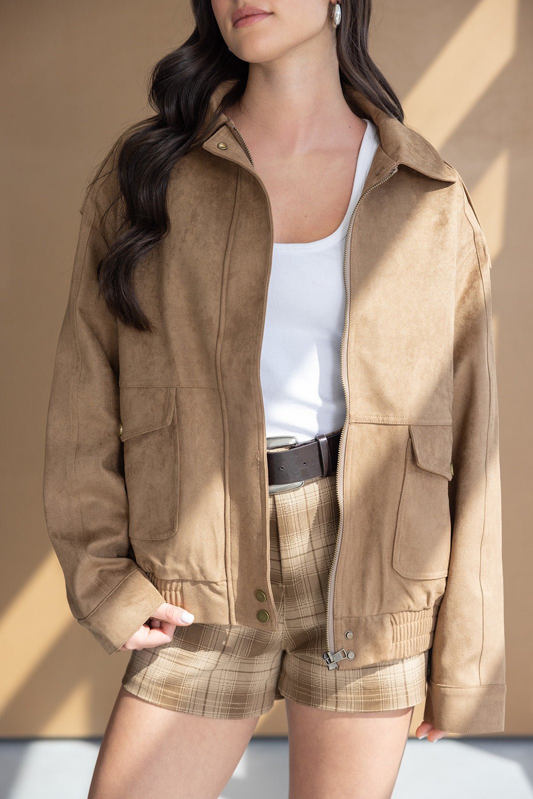 צעירה לובשת ג'קט זמש חום עליון, גופיה לבנה ומכנסיים קצרים משובצים אופנתיים