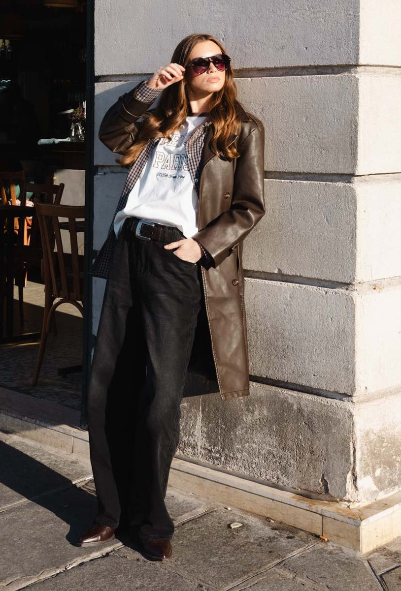 אישה מצולמת בלבוש אופנתי ברחוב, לובשת מעיל עור חום, חולצת טי PARIS, משקפי שמש ומכנסיים כהים