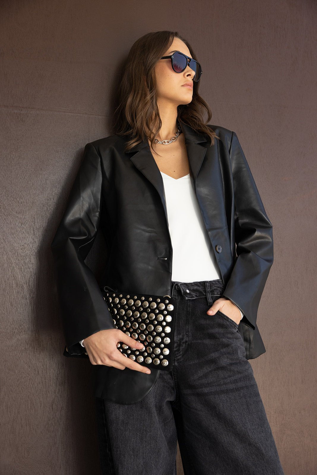 אישה עם בלייזר עור שחור, טופ לבן, מכנסי ג'ינס כהים, תיק קלאץ' עם ניטים ומשקפי שמש אופנתיות