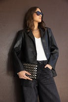 אישה עם בלייזר עור שחור, טופ לבן, מכנסי ג'ינס כהים, תיק קלאץ' עם ניטים ומשקפי שמש אופנתיות