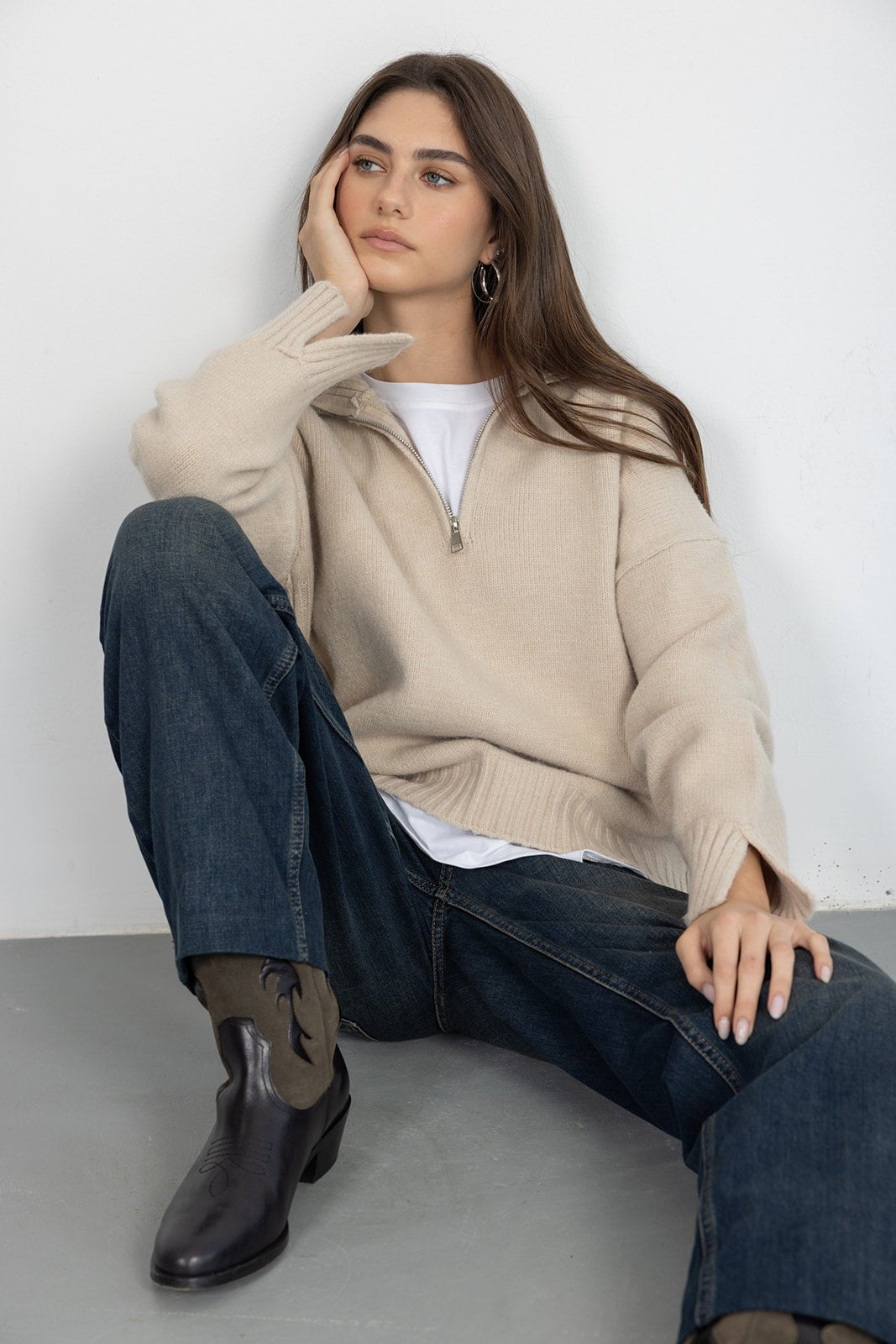 דוגמנית יושבת עם סוודר בז' רוכסן, ג’ינס ומגפיים שחורים – אופנת חורף לנשים ונערות