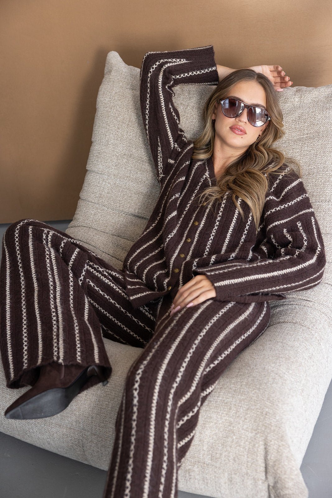 אישה יושבת על כורסה לבנה לובשת חליפת פסים חומה עם עיטורים וסוגר כפתורים, מרכיבה משקפי שמש