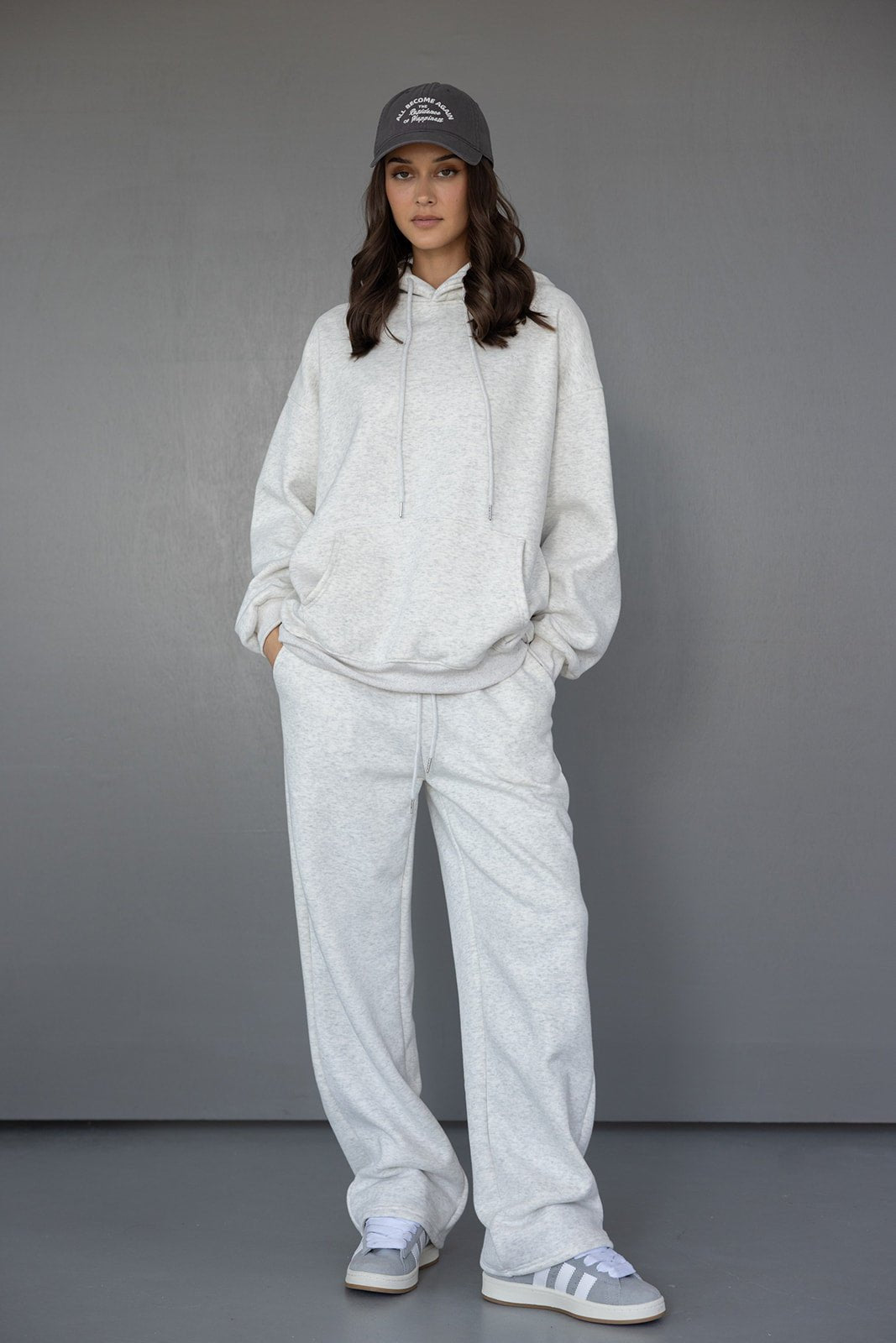 אופנת נשים: דוגמנית לובשת סט טרנינג אפור, קפוצ'ון ומכנסיים רחבים, כובע מצחייה אפור ונעלי סניקרס