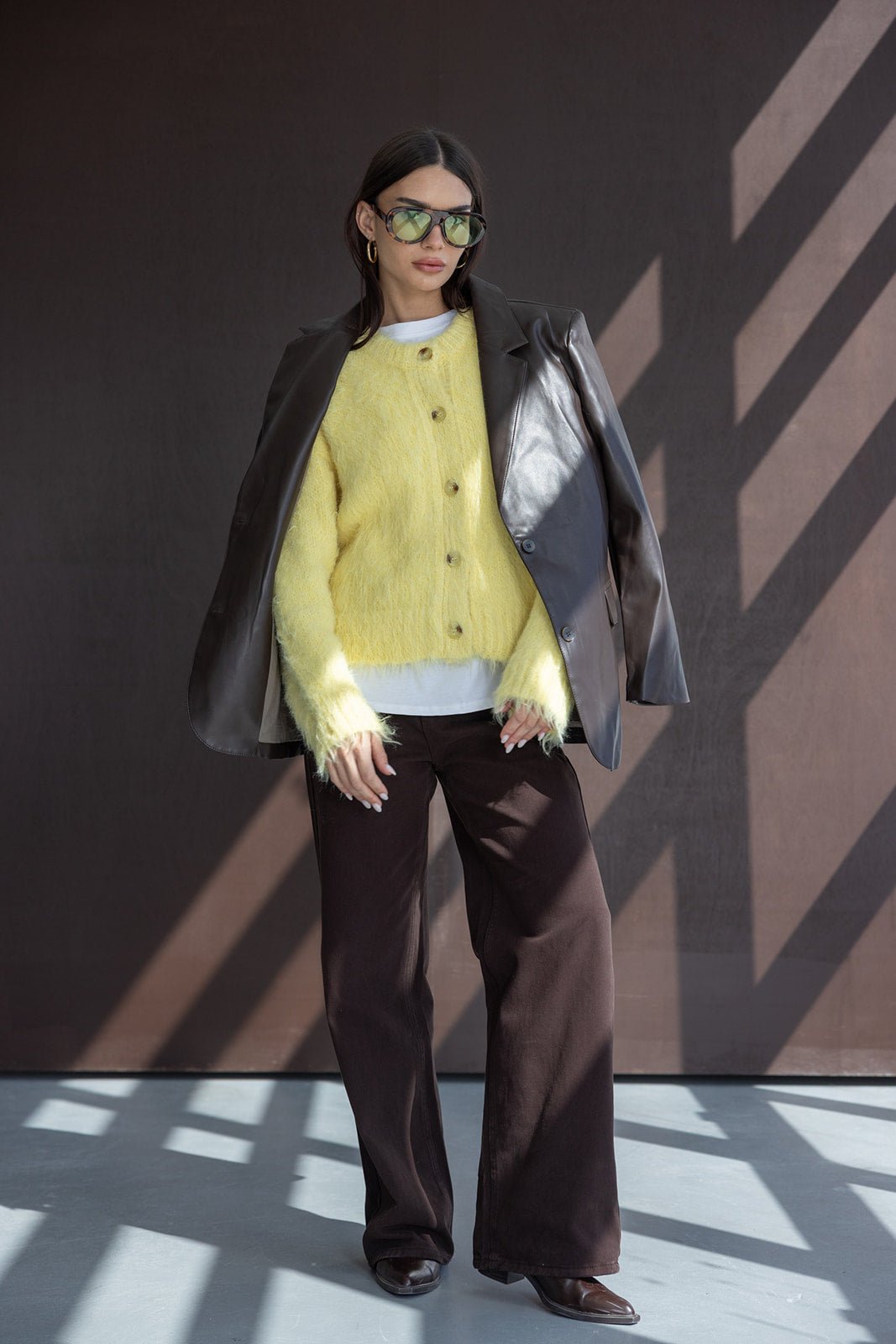 אישה עם קרדיגן צהוב פרוותי, מעיל עור חום ומשקפי שמש אופנתיים, לובשת מכנסיים רחבים בגוון כהה, סטייל אופנה לנשים