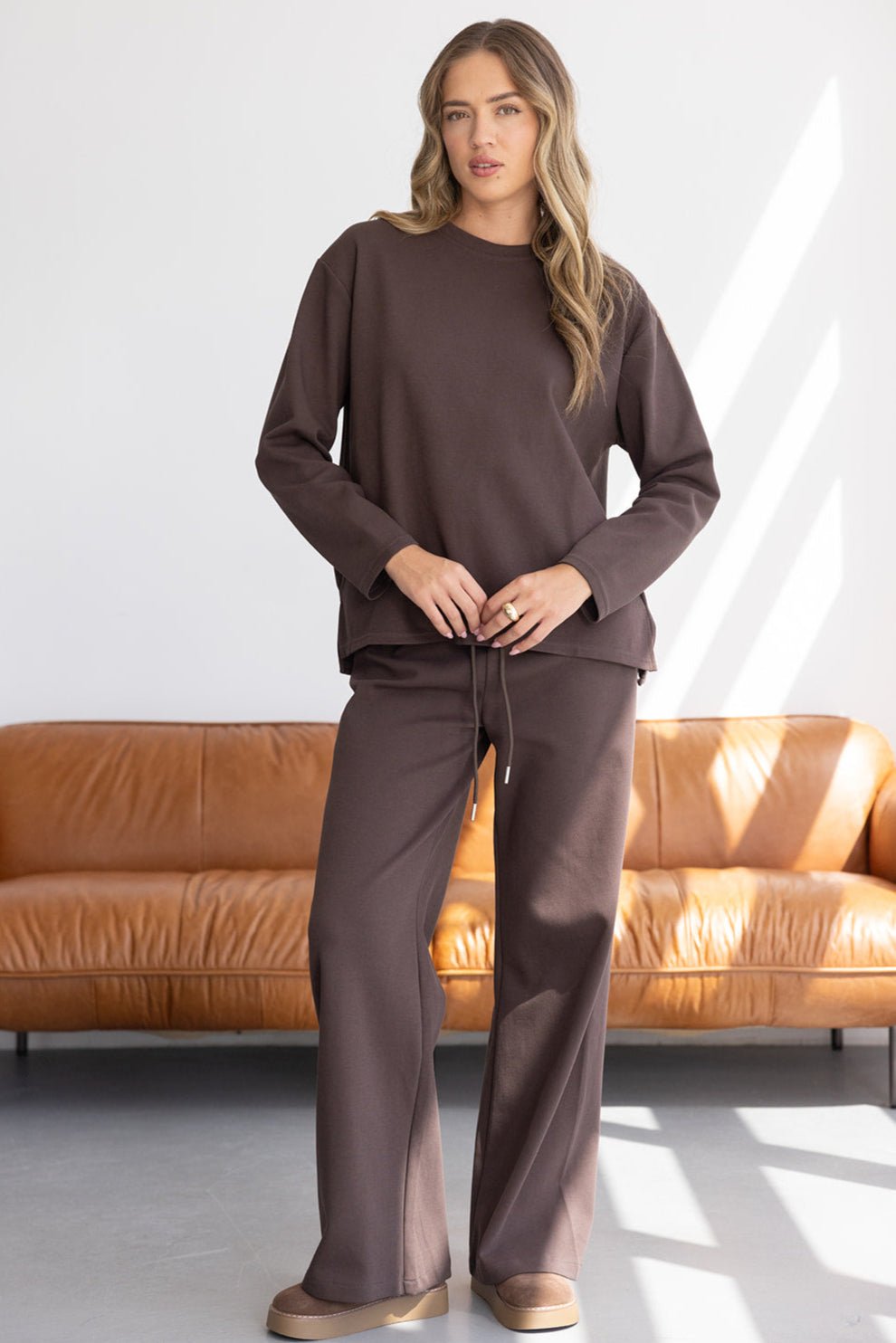 אישה לובשת סט טרנינג חום אופנתי, מכנסיים מתרחבים וסווטשירט, רקע סלון מודרני