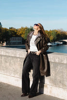 אישה אופנתית עם מעיל עור, ג'ינס רחב ומשקפי שמש עומדת על גשר בפריז ליד הנהר