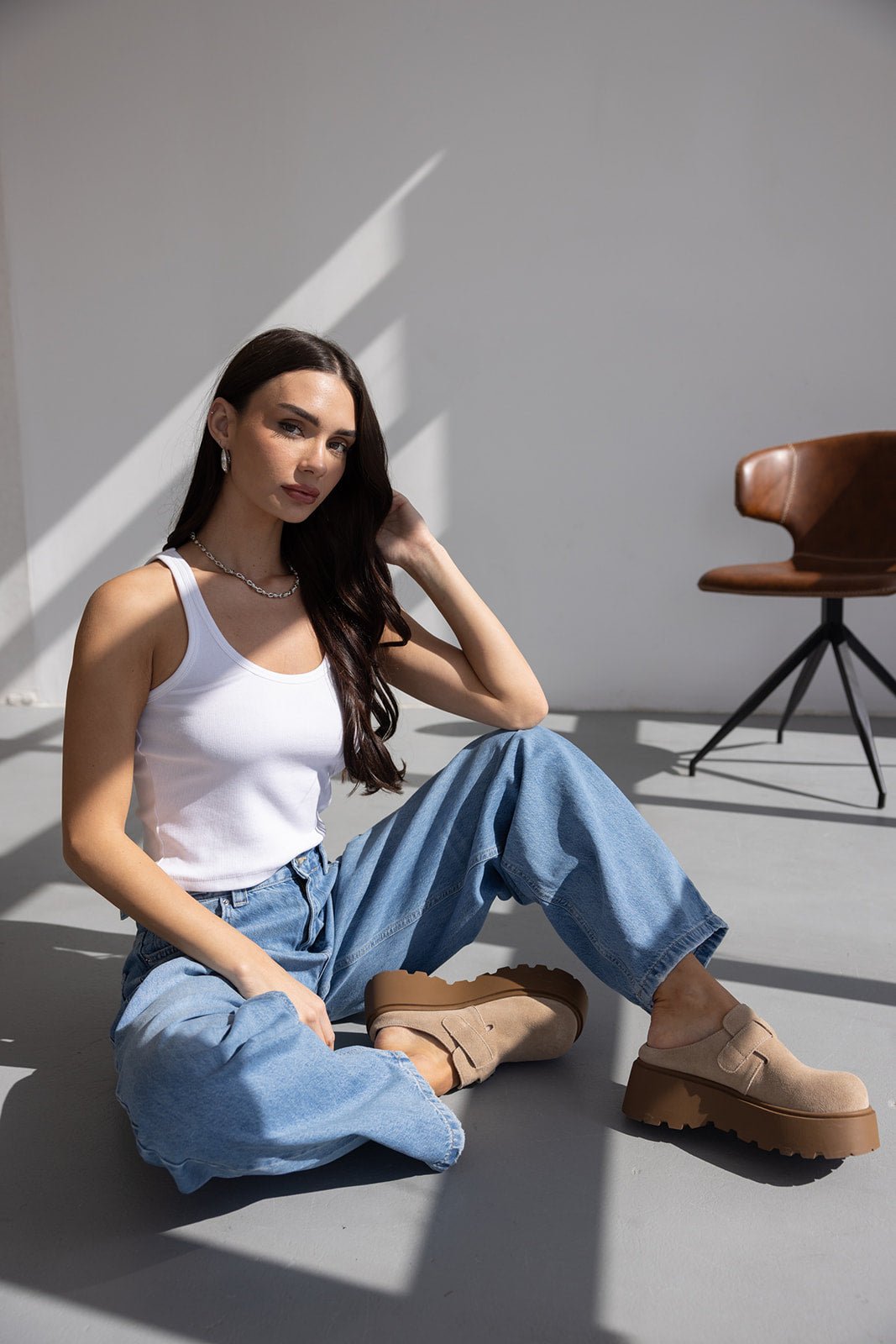 דוגמנית יושבת על רצפה בלבוש קז'ואל – גופיית טנק לבנה, ג'ינס רחב ונעלי פלטפורמה בז' אופנתיות, תאורה טבעית.