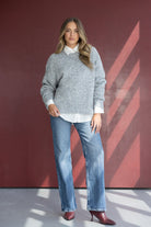 אישה לובשת סוודר אפור, חולצה לבנה, ג'ינס רחב ונעלי עקב בורדו, רקע אדום אופנתי