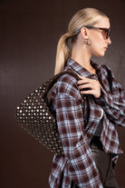 אישה עם חולצת פלאנל משובצת, תיק כתף חום עם ניטים, משקפי שמש עגולים ועגילי חישוק אופנתיים