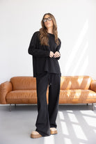 אישה צעירה עם שיער גולש לובשת סט חליפת טרנינג שחורה וצעדי פלטפורמה, עומדת על רקע ספה עור מודרנית