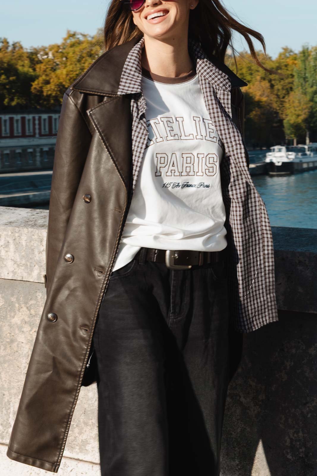 אופנת רחוב לנשים, דגם מעיל עור חום, חולצת טי שירט עם כיתוב, מכנס שחור, ברקע נהר ועצים