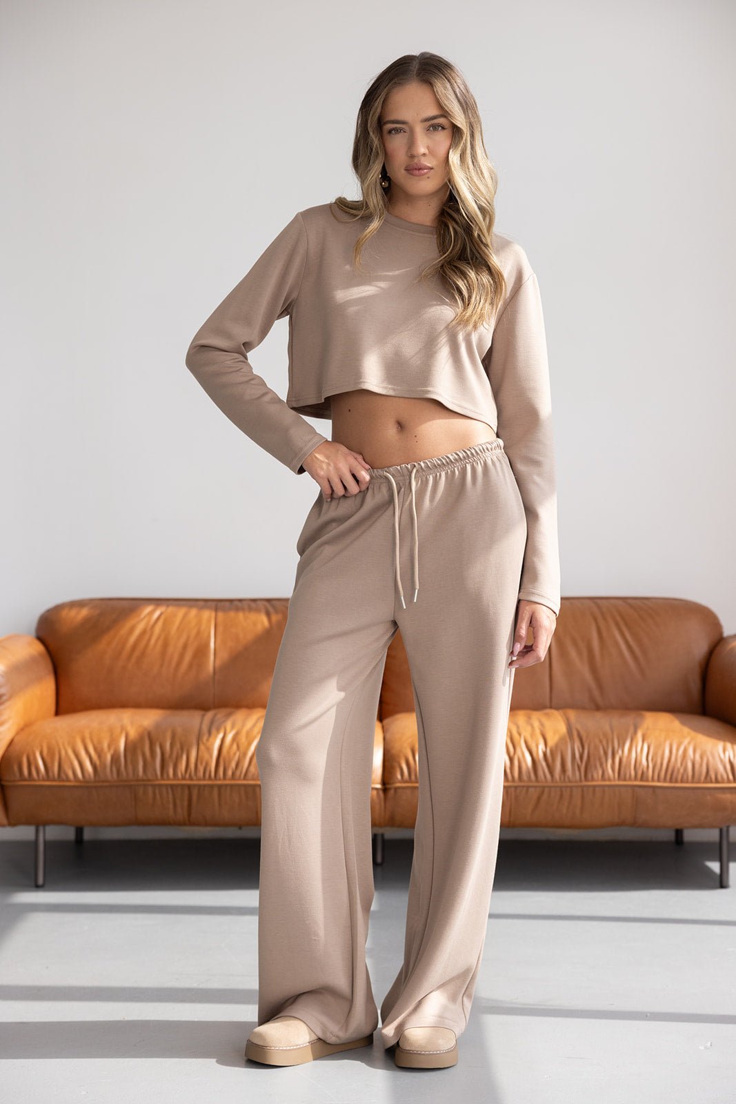 דוגמנית לובשת סט אופנה לנשים בצבע בז', חולצה ארוכה קצוצה ומכנסיים רחבים, ברקע ספה חומה
