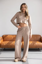 דוגמנית לובשת סט אופנה לנשים בצבע בז', חולצה ארוכה קצוצה ומכנסיים רחבים, ברקע ספה חומה