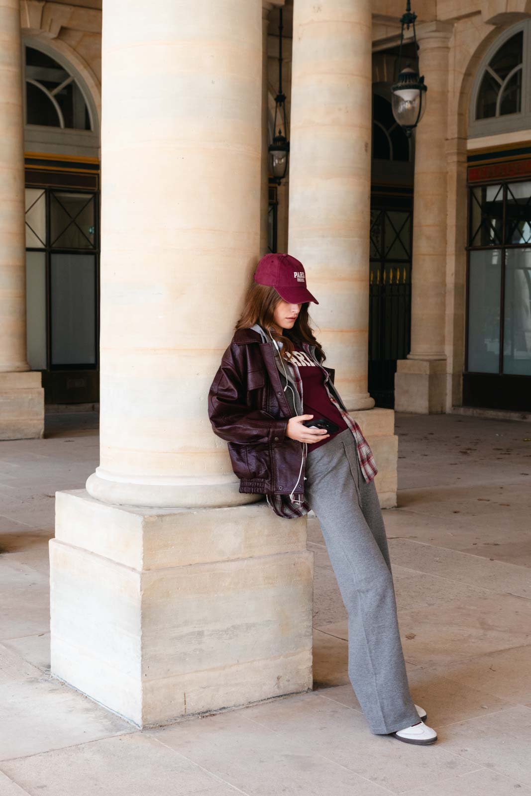 אישה צעירה עם כובע בורדו, ז'קט עור חום, חולצה אפורה ומכנסיים אפורים בלוק אופנתי בעיר