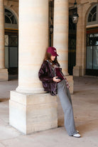 אישה צעירה עם כובע בורדו, ז'קט עור חום, חולצה אפורה ומכנסיים אפורים בלוק אופנתי בעיר