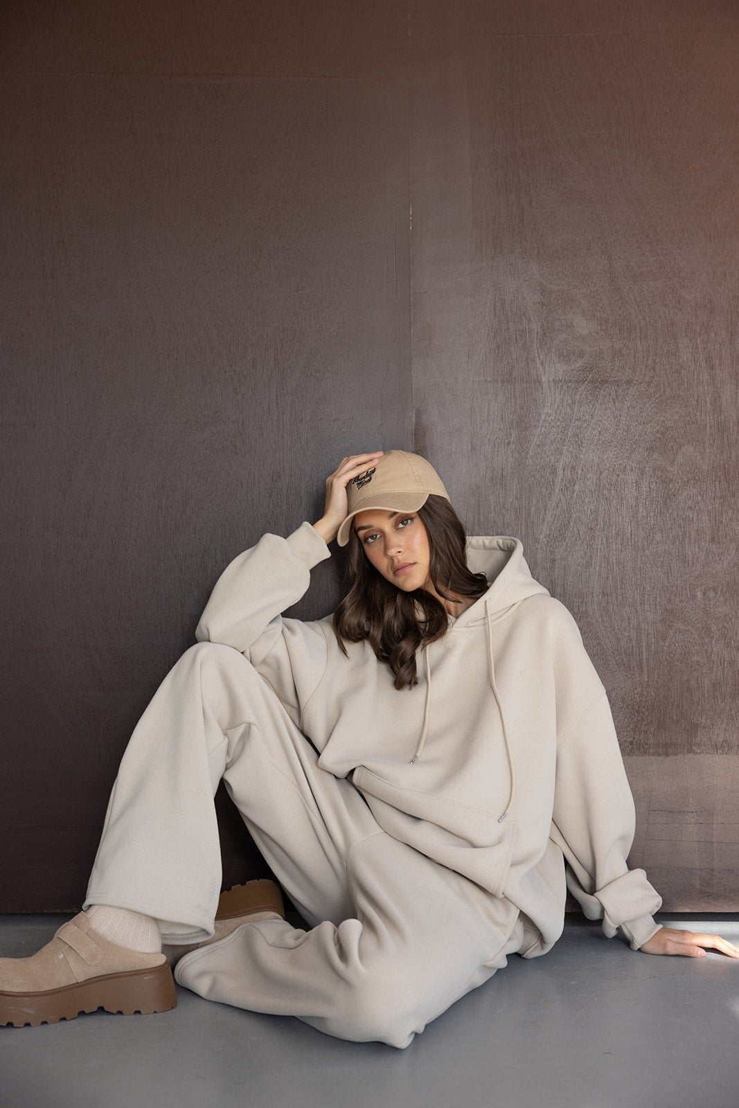 דוגמנית יושבת לובשת סט טרנינג בז', קפוצ'ון, מכנסיים וכובע אופנתי, לוק נינוח חורפי