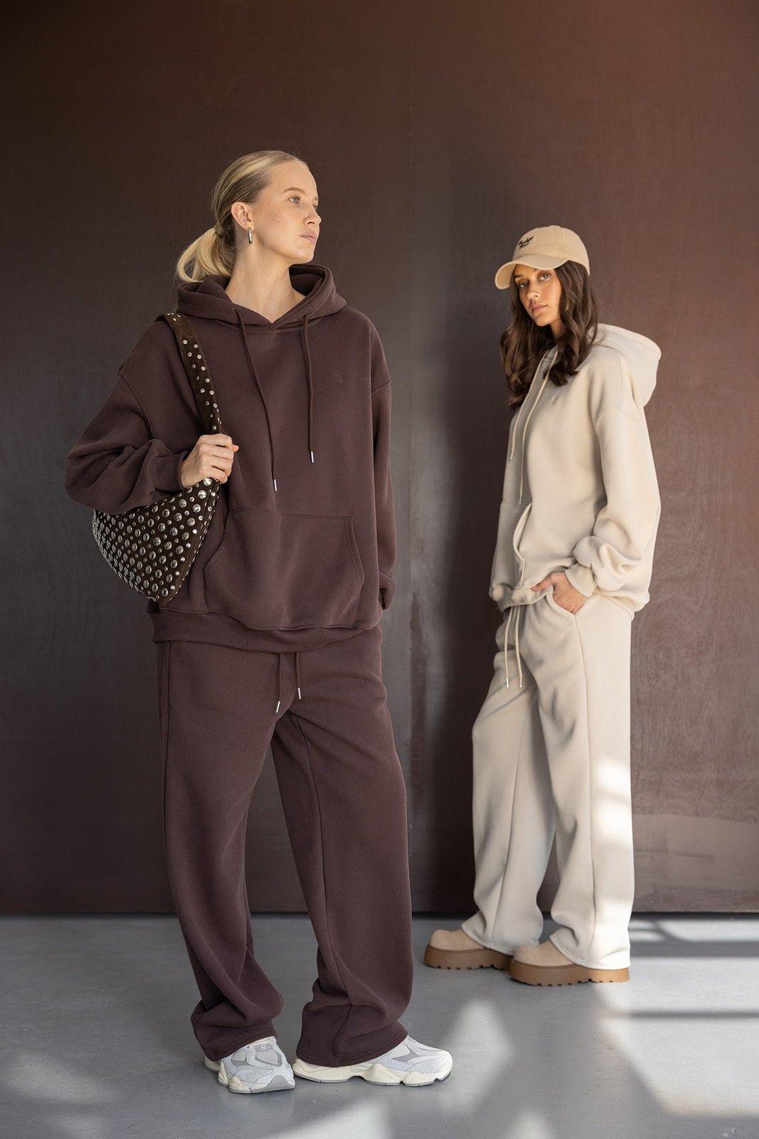 שתי נשים צעירות לובשות חליפות טרנינג אופנתיות בצבע חום ובז', חולצות קפוצ'ון ומכנסיים רחבים, אופנה לנשים וצעירות.