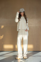 אישה עם שיער גולש חובשת כובע מצחיה, לובשת סט טרנינג נעים בגוון שמנת ונעלי בית טרנדיות, צילום אופנה ברקע נייטרלי