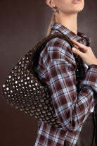 אישה עם חולצת פלנל משובצת חומה וכחולה, נושאת תיק כתף חום משובץ ניטים אופנתי