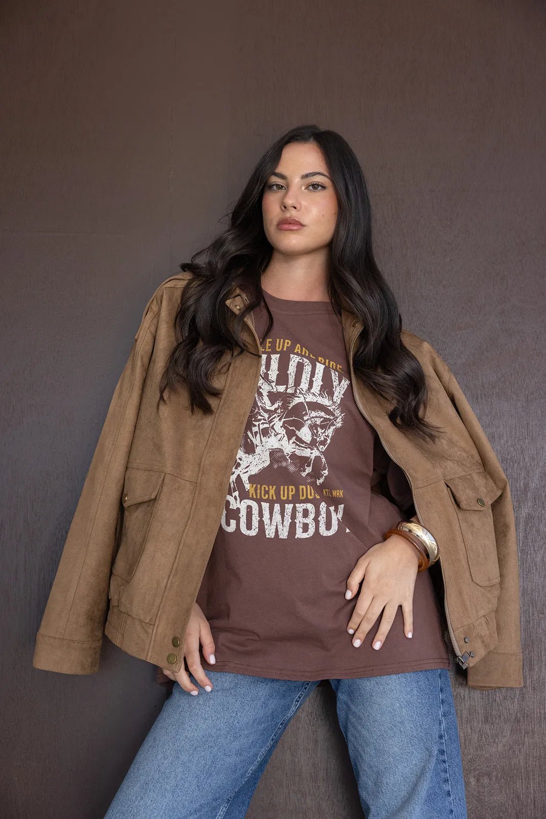 אישה צעירה לובשת טי שירט חום עם הדפס קאובוי, ג'קט זמש חום ומכנסי ג'ינס אופנתיים, אופנת נשים טרנדית.